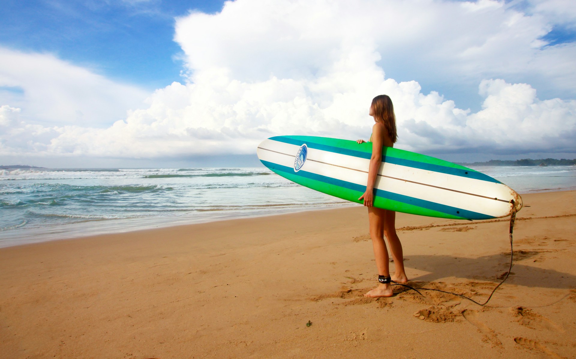 photo - a woman holding a surfboard while standing on the beach and looking towards the ocean