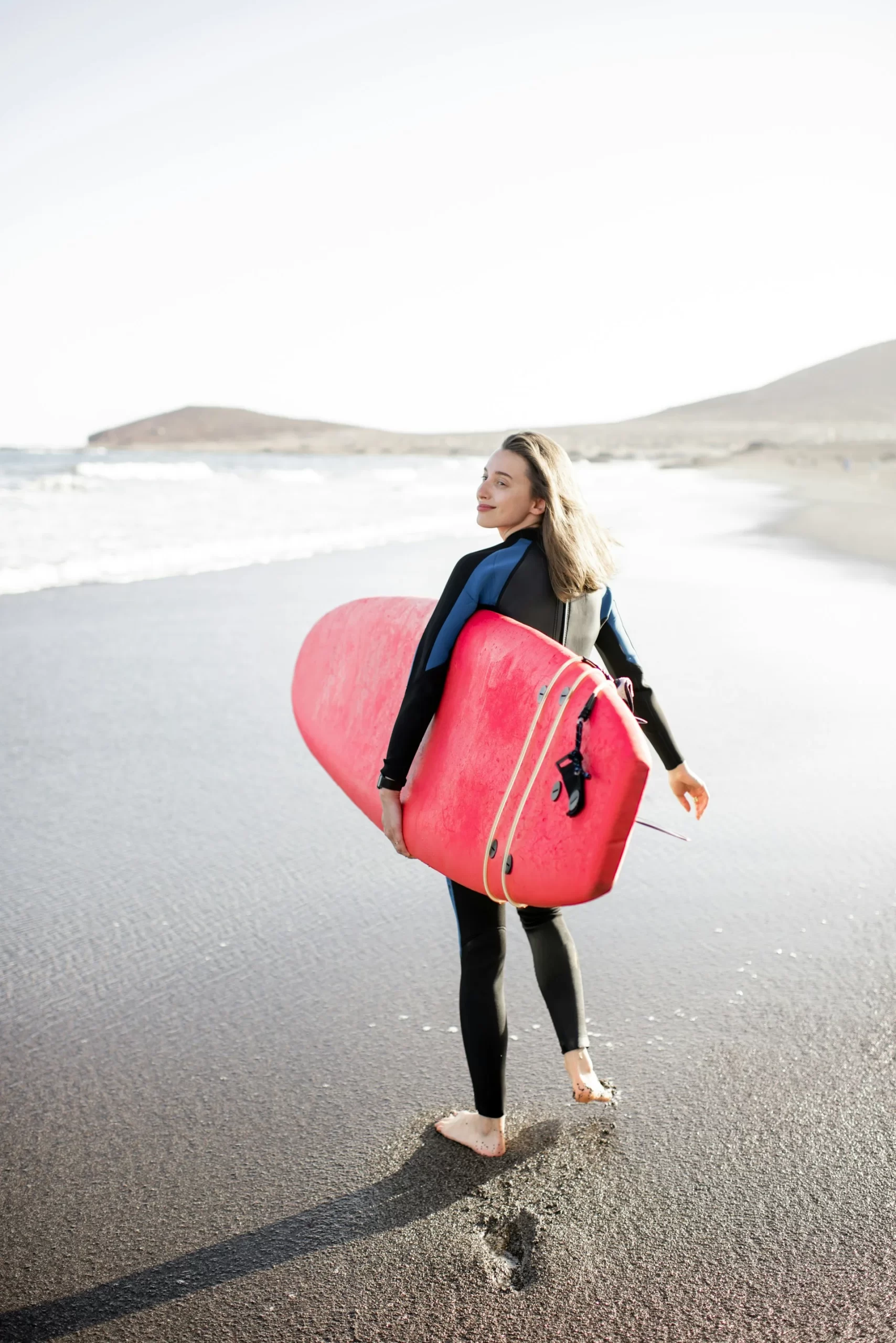photo - a woman wearing a surf hydrosuit walking down a sandy beach with a waikiki rental surf board in her hand