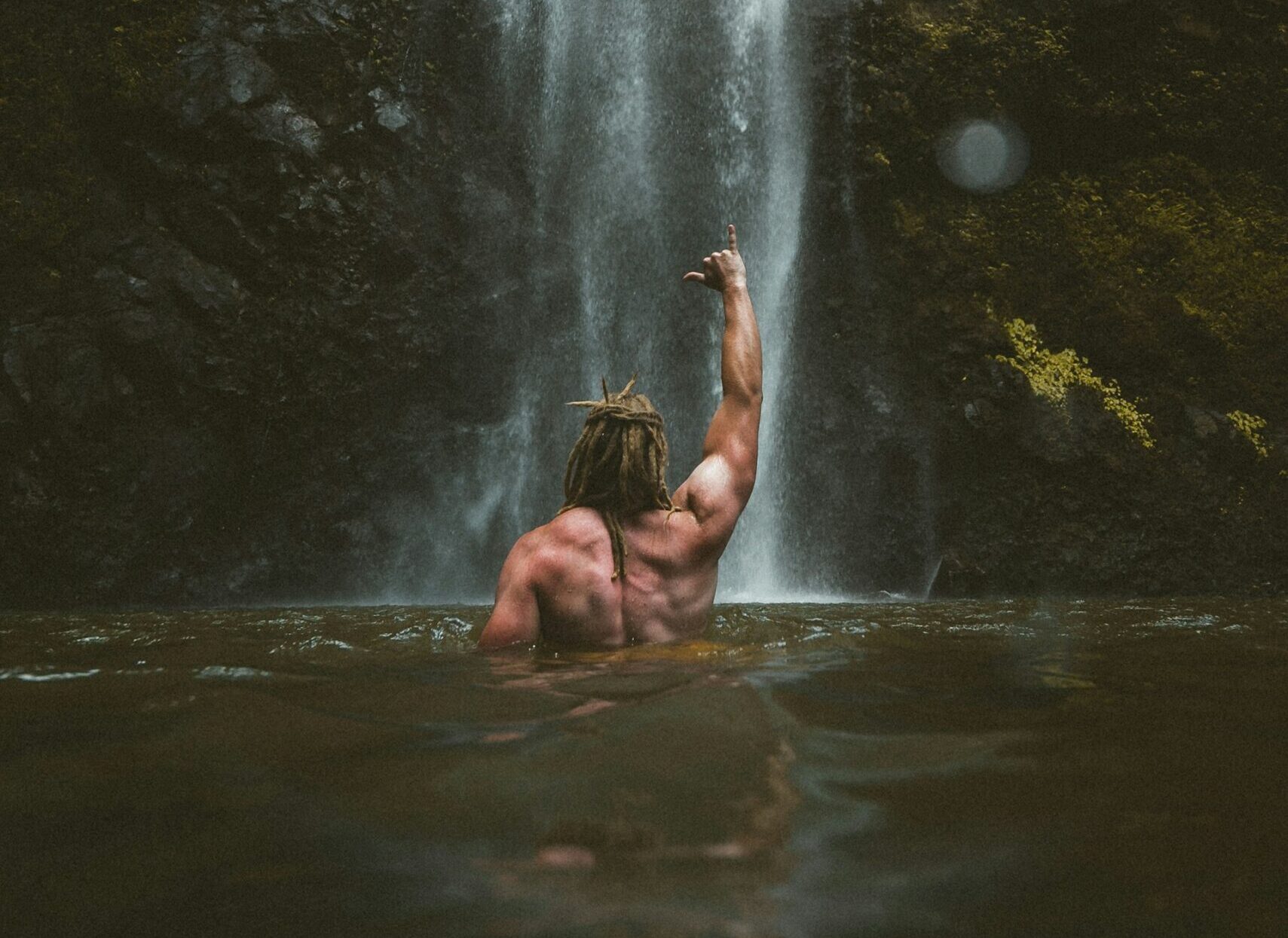 photo - a man in a waterfall lake showing a surfer sign with his hand and his back turned to the camera