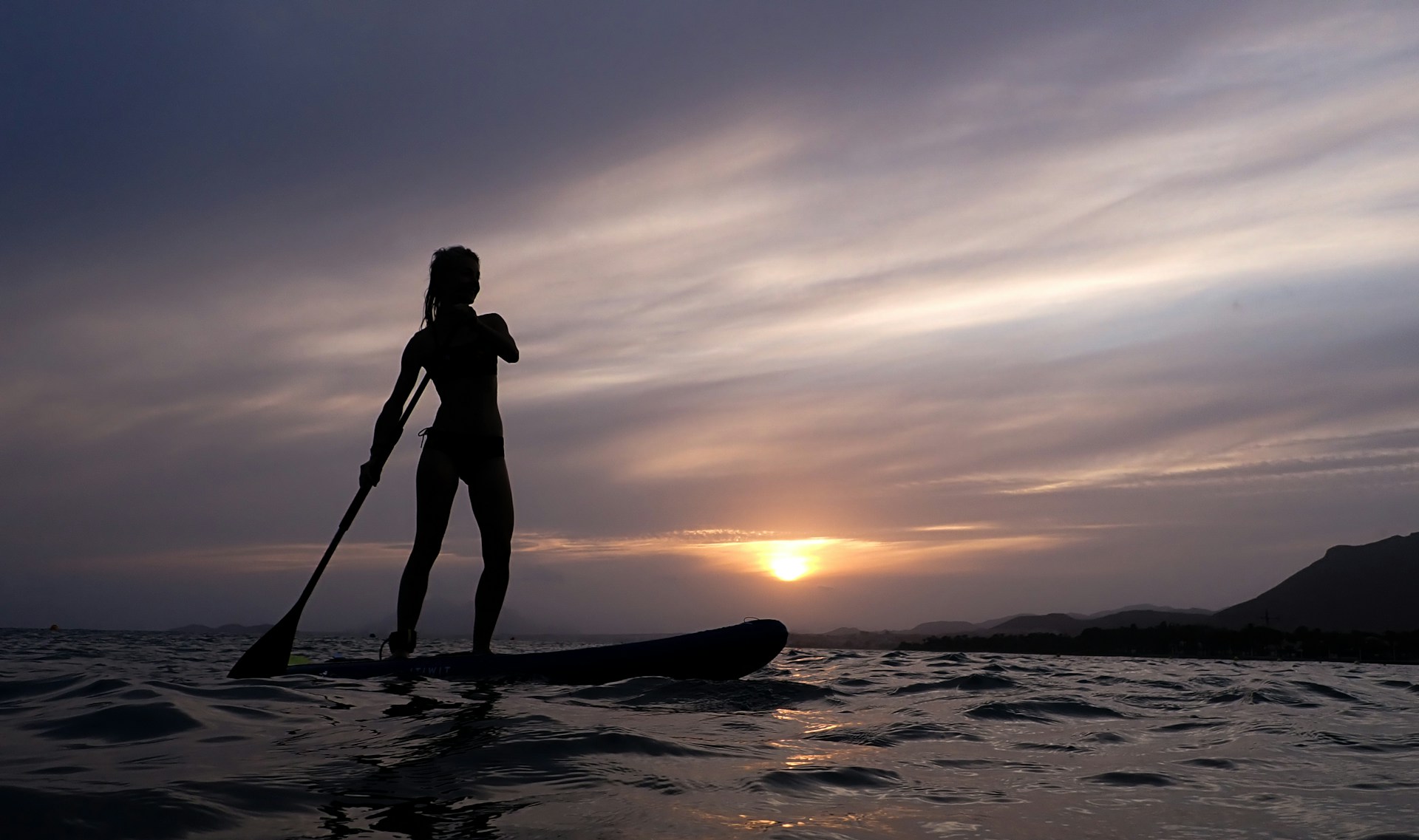 photo - a woman addle boarding in the ocean suring sunset 