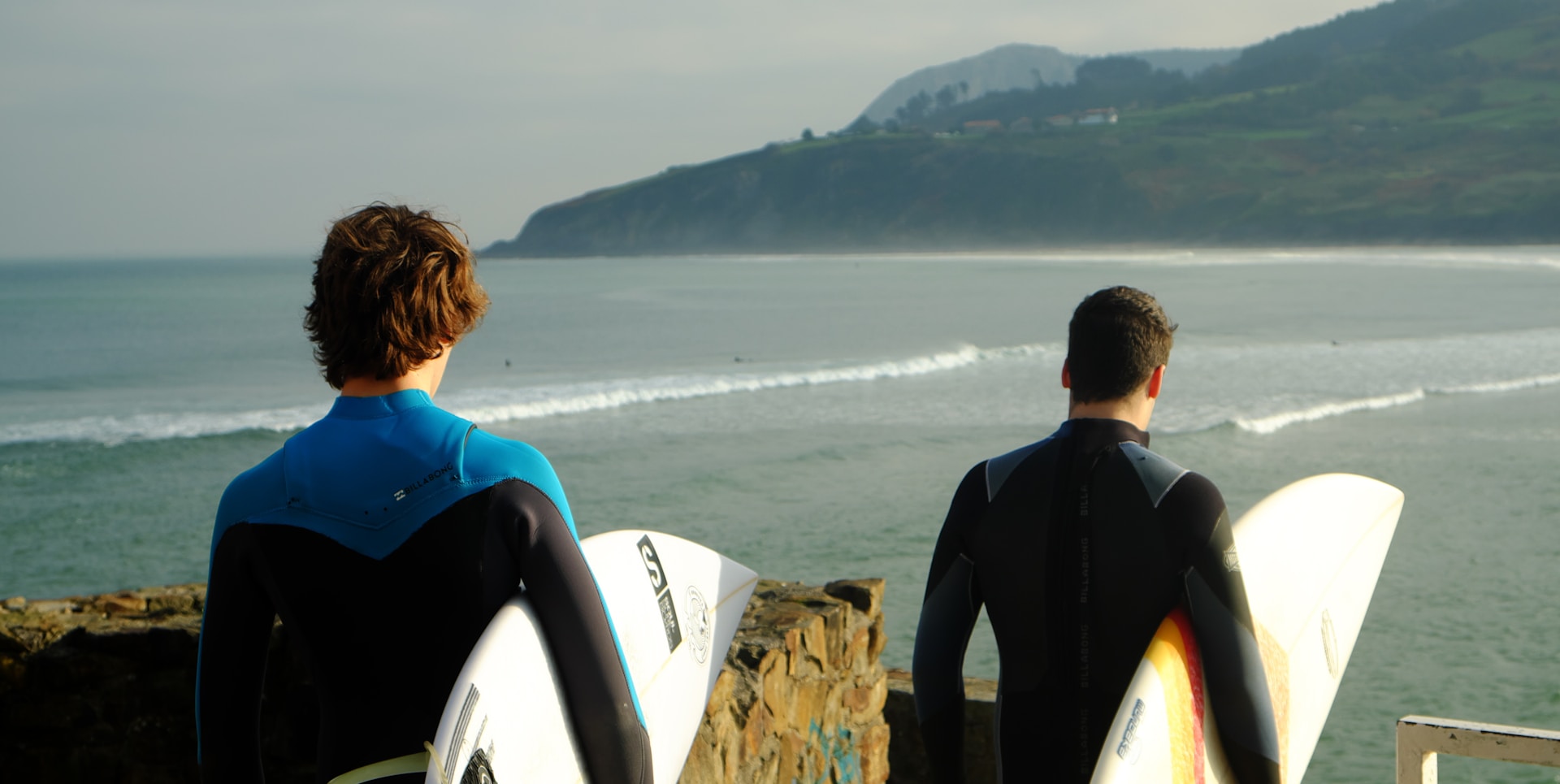 photo - two surfers walking to ride ways in a scenic location with ocean and green mountains in Hawaii