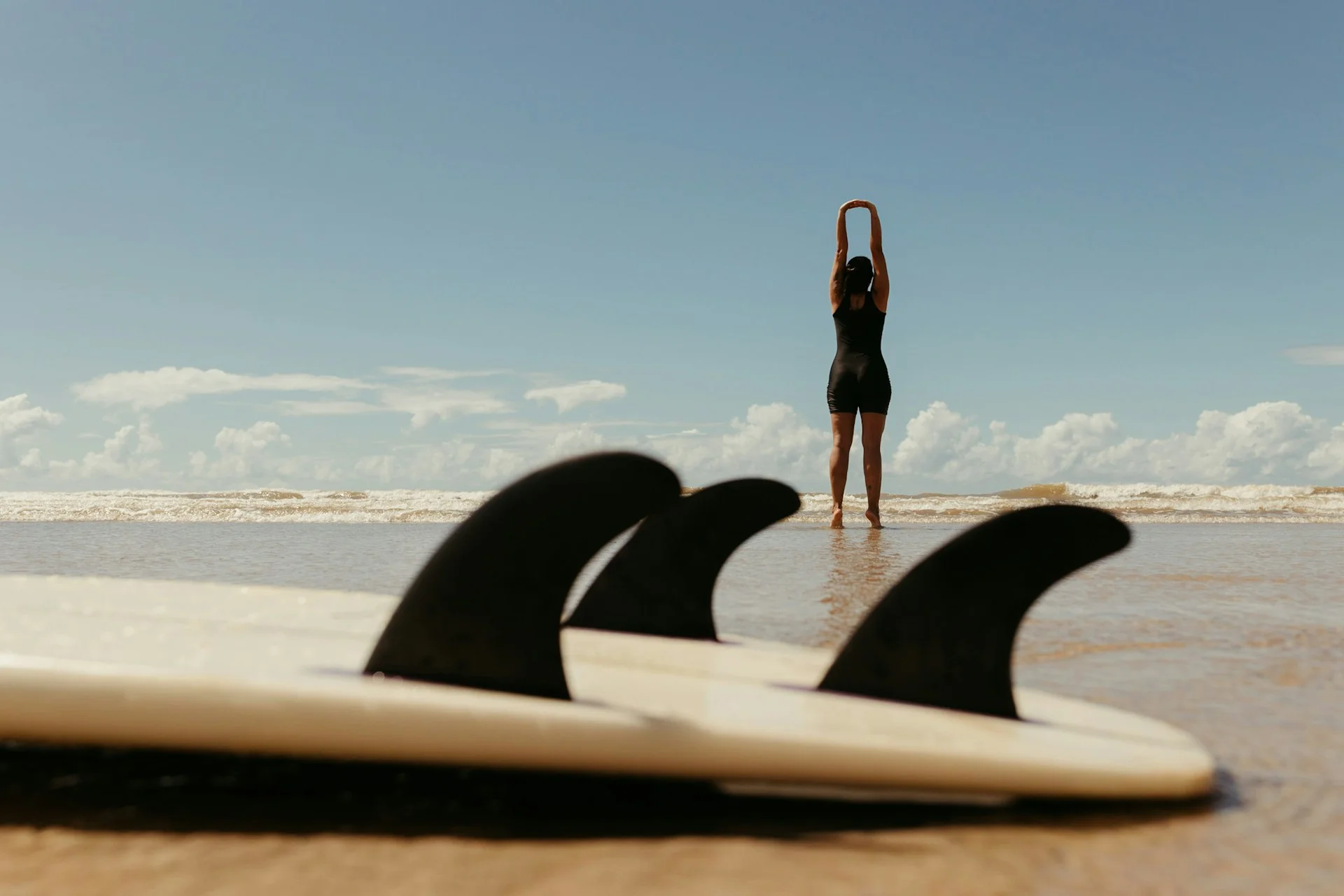 photo - a woman in the background on the beach stretching and thinking about safe surfing practices with her surfboard ont he foreground 