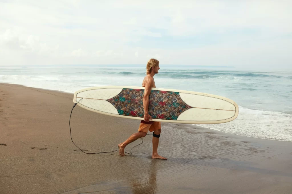 photo - a man carrying a long board to the ocean and wondering how much does a surfboard weigh