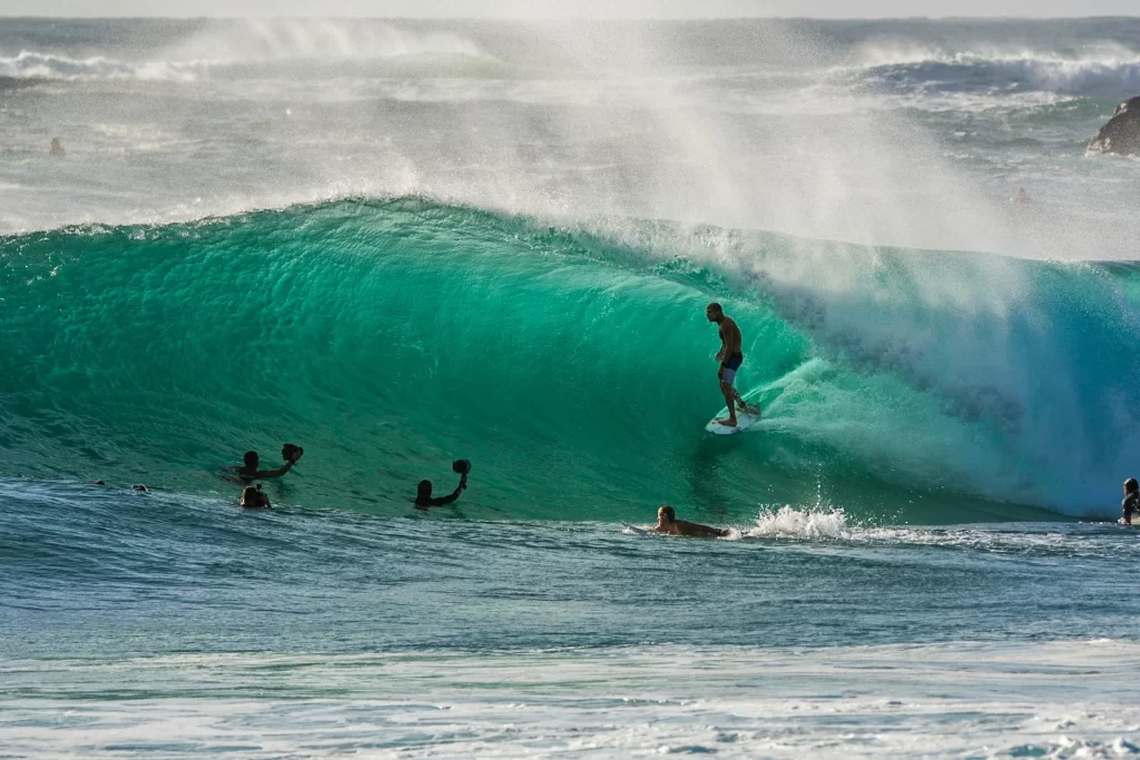 photo - a man is attempting to surf barrel with other people swimming in front of him with cameras at banzai pipeline